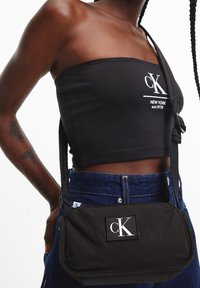 Czarna torba na ramię o prostokątnym kształcie, z widoczną, białą naszywką z logo CK, regulowanym paskiem i zapięciem na zamek.