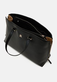 Sac à main en cuir noir avec deux poignées, logo doré sur le devant et ouverture zippée en haut révélant une doublure intérieure beige.
