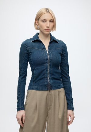 Donna con capelli biondi corti indossa una giacca di jeans blu scuro aderente con cerniera e pantaloni beige larghi su sfondo bianco semplice.