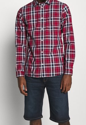 Hombre con una camisa de cuadros en rojo, blanco y negro con botones y pantalones cortos de mezclilla azul oscuro, de pie frente a un fondo liso.