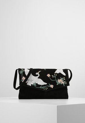 Bolso de mano de ante negro con solapa, bordado con pájaros blancos y flores rosa pastel, y una fina correa de hombro sobre una superficie blanca.
