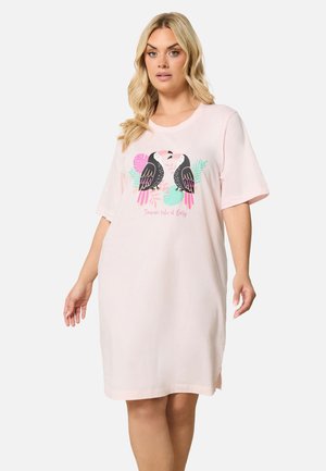 Blonde vrouw draagt een lichtroze oversized jurk met korte mouwen en een toekan afbeelding en de tekst "Toucan take it Easy" aan de voorkant.