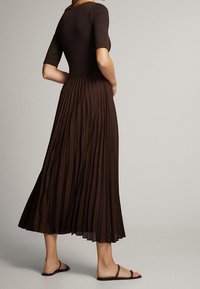 Braunes, plissiertes Kleid mit körperbetontem Oberteil und kurzen Ärmeln. Fließender Rock reicht bis zu den Kn öcheln. Getragen mit flachen Sandalen.