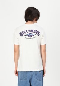 Off-white t-skjorte med korte ermer som har "BILLABONG" i lilla grafisk skrift og bølgedesign på baksiden, laget av bomull.