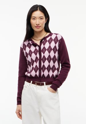 Frau trägt eine lila und pinke Argyle-Strickjacke, weiße Hose mit braunem Gürtel und eine goldene Kette, steht mit einer Hand in der Tasche.