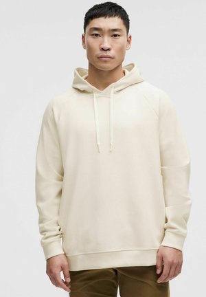 Beige hooded sweatshirt with a smooth texture, raglan sleeves, and drawstrings. Minimalist design, paired with brown trousers.