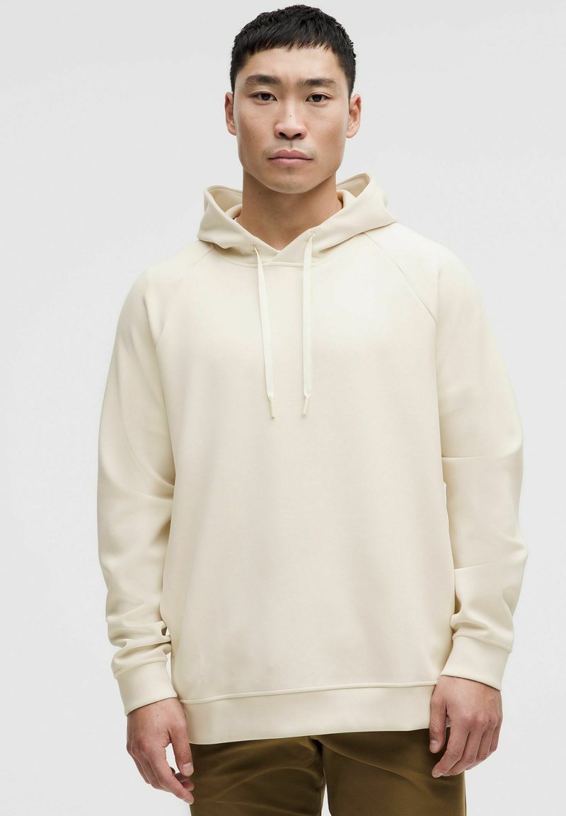 Hanorac bej cu glugă, cu o textură netedă, mâneci raglan și șnururi. Design minimalist, asortat cu pantaloni maro.