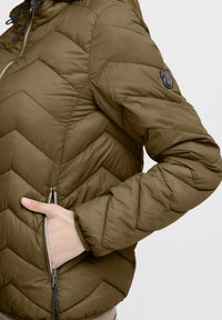 Alyvuogių žalios spalvos pamušta striukė su zigzago raštu, kišenėmis su užtrauktukais ir logotipo antspaudu ant rankovės. Lygus, blizgus audinys.