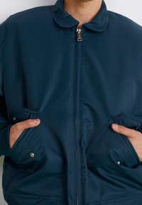 Giacca isolante blu navy con zip frontale, colletto arrotondato e tasche laterali. Texture liscia con cuciture visibili e accenti metallici.