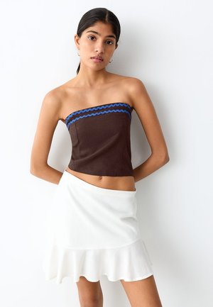 Vrouw draagt een strapless bruin topje met blauwe bies en een witte, gerimpelde minirok, staand tegen een witte muur met handen achter de rug.