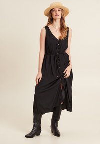 Zwarte maxi-jurk met knopen en een strik in de taille, V-hals en split aan de zijkant, gecombineerd met zwarte cowboylaarzen en een strohoed.