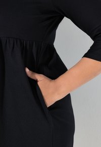 Černé šaty s přiléhavým živůtkem a nařasenou sukní. Mají postranní kapsu a tříčtvrteční rukávy, vyrobené z hladké elastické látky.