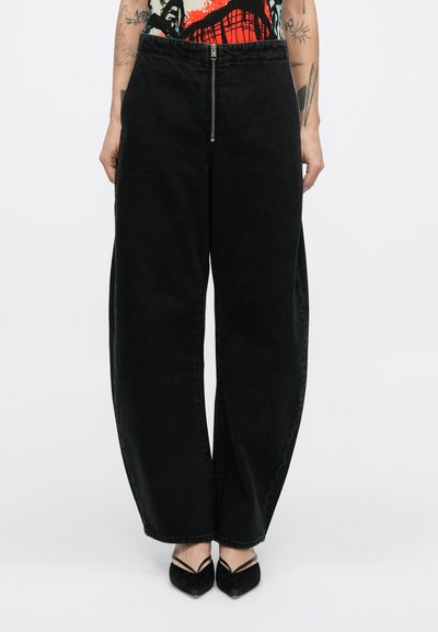 Pantaloni neri in denim a gamba larga con vita alta, caratterizzati da una cerniera frontale e una texture sottile. Indossati con scarpe nere a punta.
