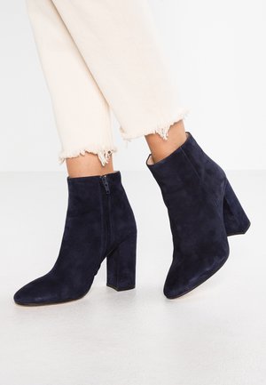 Marineblaue Wildleder-Stiefeletten mit spitzer Zehenpartie, Reißverschluss an der Seite und klobigem Absatz. Getragen mit beigefarbenen, gekürzten Hosen mit ausgefransten Säumen.