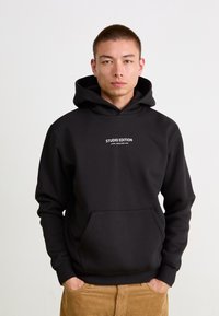 Felpa nera di tessuto morbido con tasca frontale, cappuccio regolabile e testo bianco che recita "STUDIO EDITION ALPHA INDUSTRIES INC."