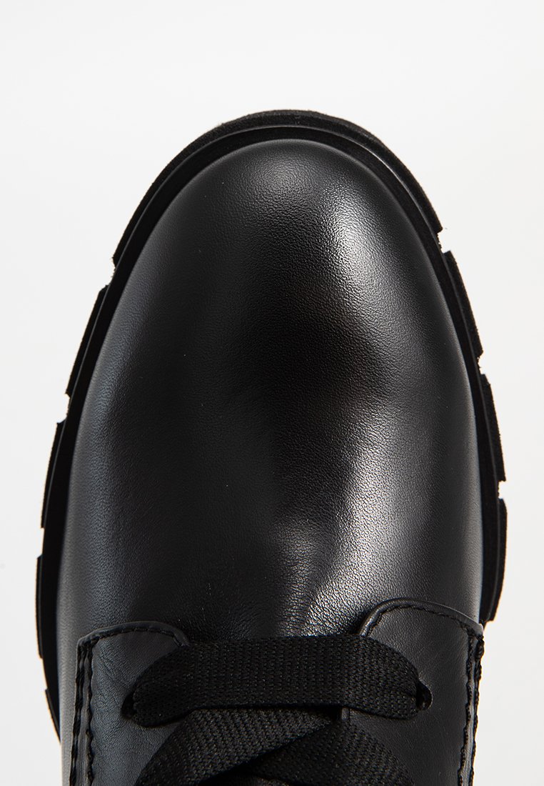 Gros plan sur l'embout arrondi d'une botte en cuir noir avec des bords de semelle texturés et des lacets en tissu noir sur fond blanc.