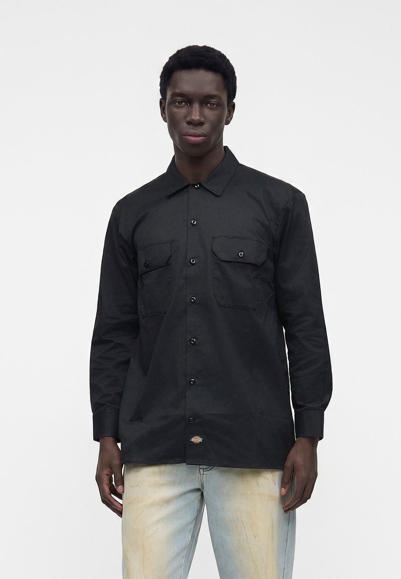 Mand iført en sort, knappet langærmet skjorte med to brystlommer og lyseblå, falmede jeans, stående foran en ensfarvet baggrund.