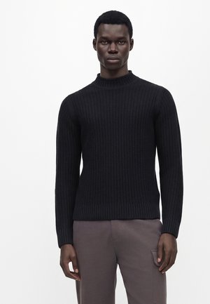 Jeune homme portant un pull en maille côtelée noire et un pantalon marron, debout devant un fond clair uni.
