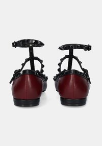 Bailarinas de cuero rojo con correas de tobillo de charol negro, con tachuelas en forma de pirámide y diseño de punta redondeada. Textura suave y herrajes minimalistas.