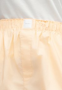 Ελαστική μέση σε ανοιχτό μπεζ σορτς Calvin Klein, φορεμένα με λευκό πουκάμισο, με έμφαση στην υφή του υφάσματος και την ετικέτα της μάρκας.