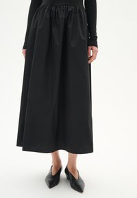 Fustă midi neagră realizată dintr-un material neted, cu un talie plisată. Puțină cu pantofi cu toc ascuțit negri. Siluetă simplă și elegantă.