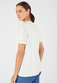 Top corto bianco a maniche corte con ricamo a occhio di pernice sulle maniche a sbuffo. Tessuto morbido con un fit rilassato, orlo arrotondato e design minimalista.