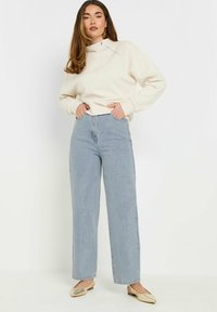 Felpa crema con collo alto e coulisse, abbinata a jeans a vita alta e gamba larga di un blu chiaro. Scarpe slip-on dorate completano l'outfit.