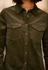 Camicia in corduroy verde scuro con chiusura a bottoni, due tasche sul petto e un colletto classico con bottoni a pressione argentati.