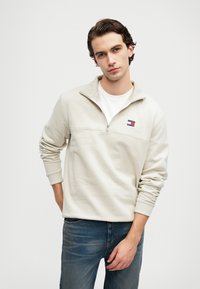 Jasnobeżowy pulower z kołnierzykiem na zamek, z małym, wielokolorowym logo na piersi. W połączeniu z niebieskimi dżinsami. Miękka, strukturalna tkanina.