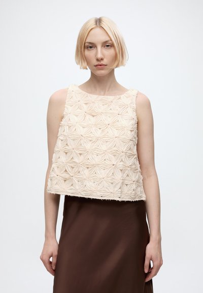 Femme aux cheveux blonds courts portant un haut sans manches crème texturé avec des motifs floraux et une jupe marron, debout devant un fond uni.