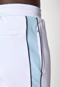 Λευκά αθλητικά παντελόνια με μπλε διχτυωτό πάνελ και μαύρες ρίγες. Περιλαμβάνει πλαϊνή τσέπη με φερμουάρ και ελαστική μέση για εφαρμοστό στυλ.