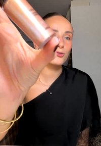 Donna con top nero che tiene vicino alla telecamera un tubetto di prodotto make-up, dimostrandolo o recensendolo all'interno.