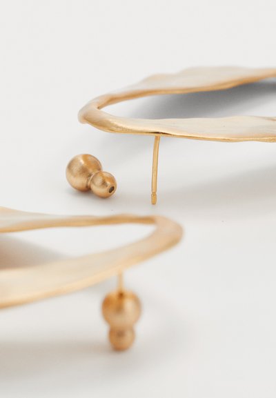 Two irregular-shaped gold hoop earrings with small round gold backings on a white surface.