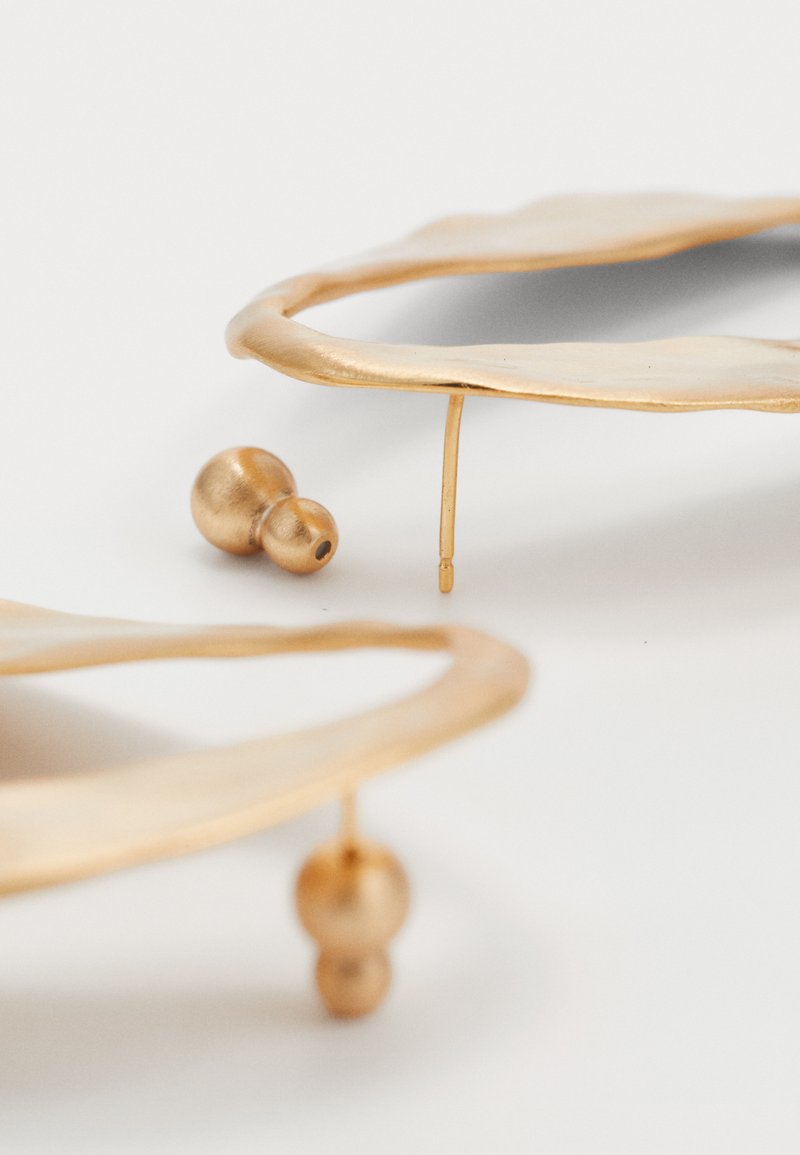 Two irregular-shaped gold hoop earrings with small round gold backings on a white surface.