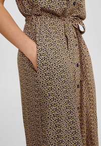 Vestito con stampa leopardata su base beige, caratterizzato da macchie nere e gialle, cintura in vita, tasche laterali e design con bottoni.