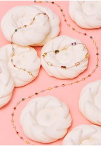 Gold chain jewelry featuring multicolored gem accents, arranged atop white meringue cookies, against a pink background.