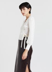 Cardigan bianco lavorato a maglia con motivo a diamante, cintura in vita, e maniche lunghe; abbinato a una gonna marrone scuro con spacco alto.