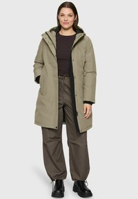 Vrouw in een beige winterjas met capuchon, een donkerbruin overhemd met lange mouwen, losse bruine broek en zwarte enkelboots, staand tegen een effen achtergrond.