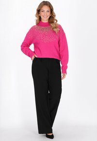 Jasnoróżowy sweter z dzianiny z wysokim kołnierzem, zdobiony srebrnymi elementami ozdobnymi na górnej części klatki piersiowej, w połączeniu z czarnymi spodniami o szerokich nogawkach.
