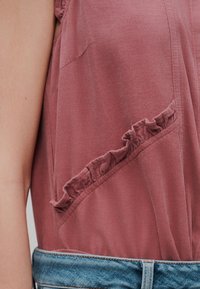 Haut sans manches en velours côtelé rose avec un détail de bordure à volants le long d'une couture diagonale, porté avec la ceinture visible d'un jean en denim bleu.