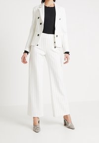 Valkoraitainen bleiseri mustilla napeilla, yhdistettynä mustaan ribbipaitaan ja leveälahkeisiin valkoisiin housuihin. Korostettu hopeanvärisillä käärmeennahkakuosisilla kenkillä.