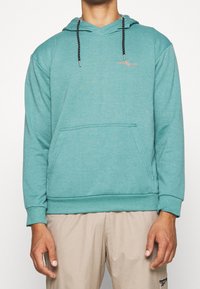 Felpa color teal con tasca frontale, cappuccio con cordini e accenti neri a contrasto. Realizzata in tessuto morbido con vestibilità comoda.