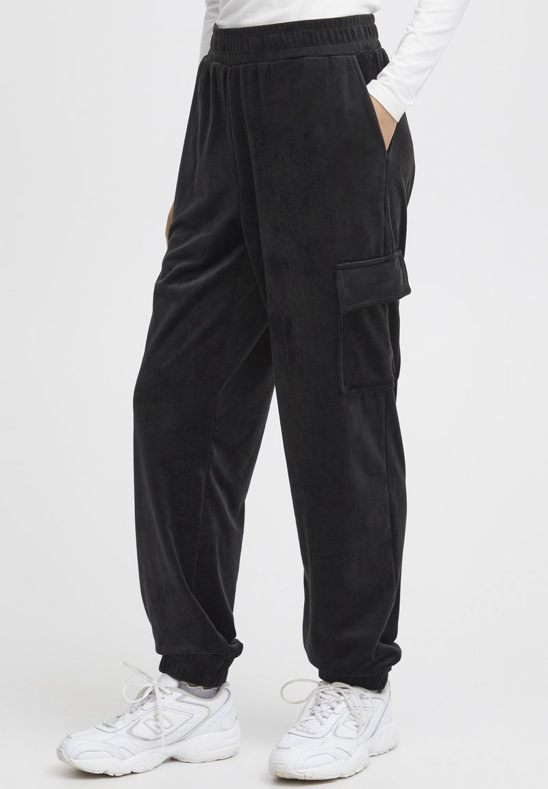 Zwarte fleece joggers met een elastische tailleband, zijzakken en een cargozak. De boorden zijn elastisch. Gemaakt in combinatie met witte sportschoenen.