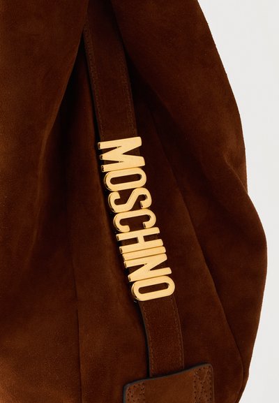 Καφέ τσάντα από σουέντ με ιμάντα που φέρει μεγάλα χρυσά γράμματα που σχηματίζουν τη λέξη "MOSCHINO" κατακόρυφα.