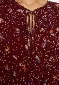 Blusa floreale bordeaux con scollatura a V, dettaglio con fiocco al collo e motivi floreali bianchi, blu e arancioni sparsi in tutto il capo.