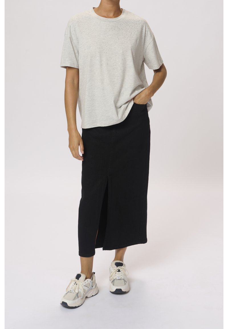 Personne portant une chemise à manches courtes gris clair, une jupe midi noire fendue à l'avant et des baskets blanches, debout avec une main dans la poche.