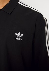 Musta polo-paita, jossa valkoinen Adidas-logo vasemmassa rinnassa, olkapäillä valkoiset raidat ja klassinen kaulusdesign.