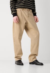 Pantalon en coton beige avec une coupe décontractée et un design classique à cinq poches, assorti à des chaussures en cuir noir et un pull rayé noir et olive.