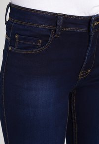 Mörkblå denimjeans med gula sömmar, som har en framficka, slät textur och en figurnära design. Högmidjade med knäppning.