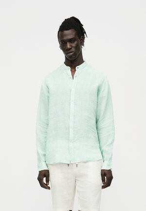 Homme à la peau foncée et aux dreadlocks porte une chemise boutonnée vert clair à manches longues et un pantalon blanc sur un fond clair.